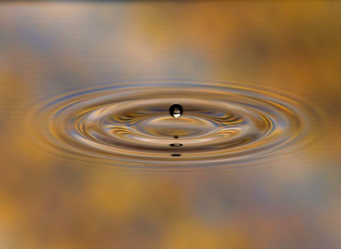 A small drop bouncing above concentric ripples on a liquid bath, lit in gold and blue.