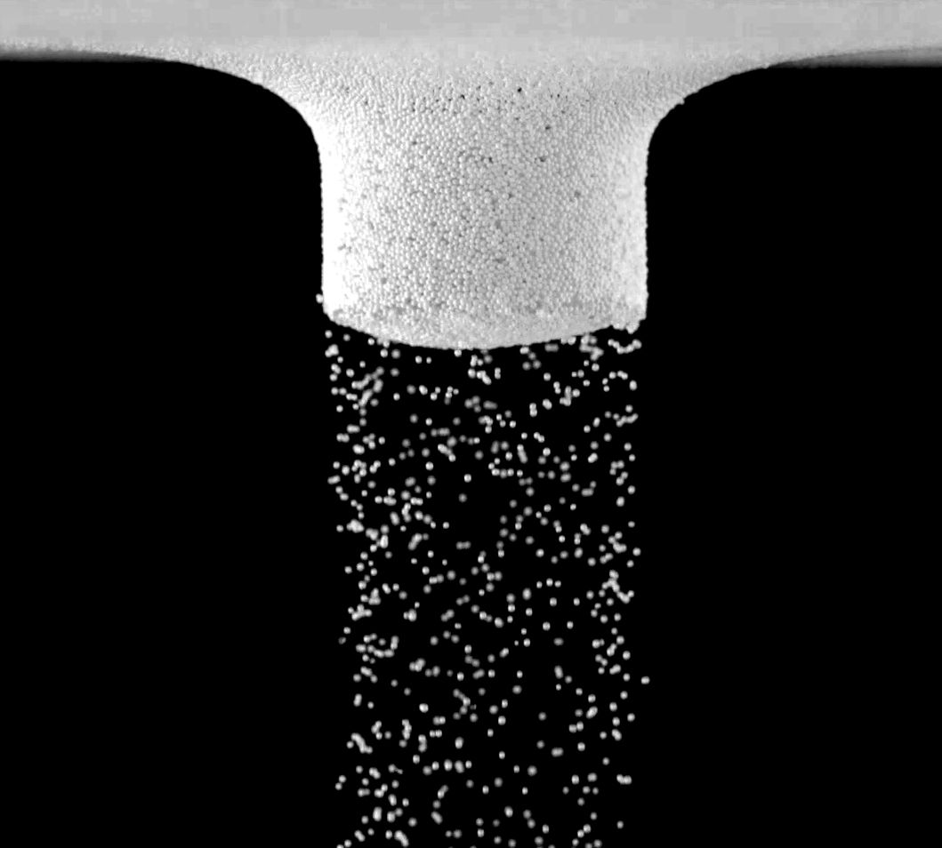 A particle-coated cylinder face with a stream of white particles falling away below it, on black.